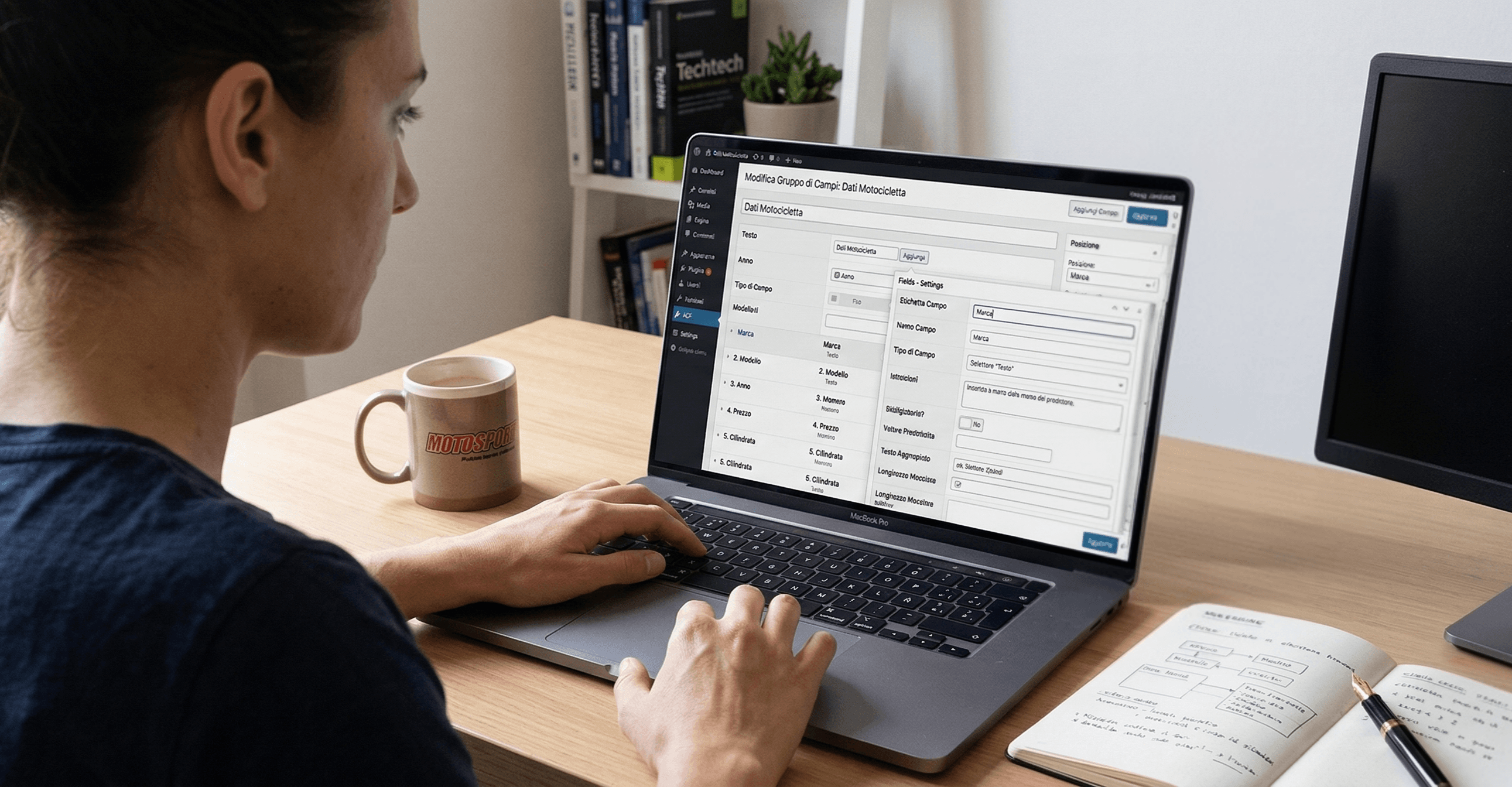 Una sviluppatrice di fronte a un macbook portatile con l'interfaccia di Wordpress e ACF, presenti un quadernetto, una tazza e un monitor